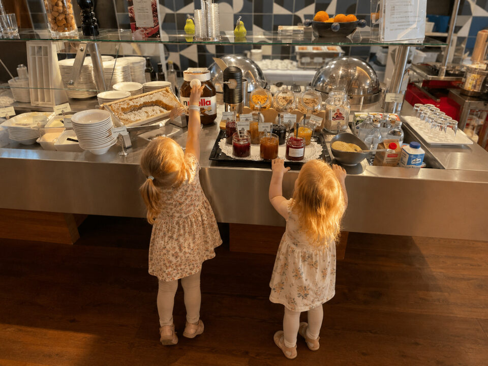 Zwei Kinder strecken sich nach einem Nutella-Glas am Frühstücksbuffet im Hotel Ploberger in Wels. Ein liebevoller Moment voller Vorfreude auf das gemeinsame Frühstück im Morgenlicht.